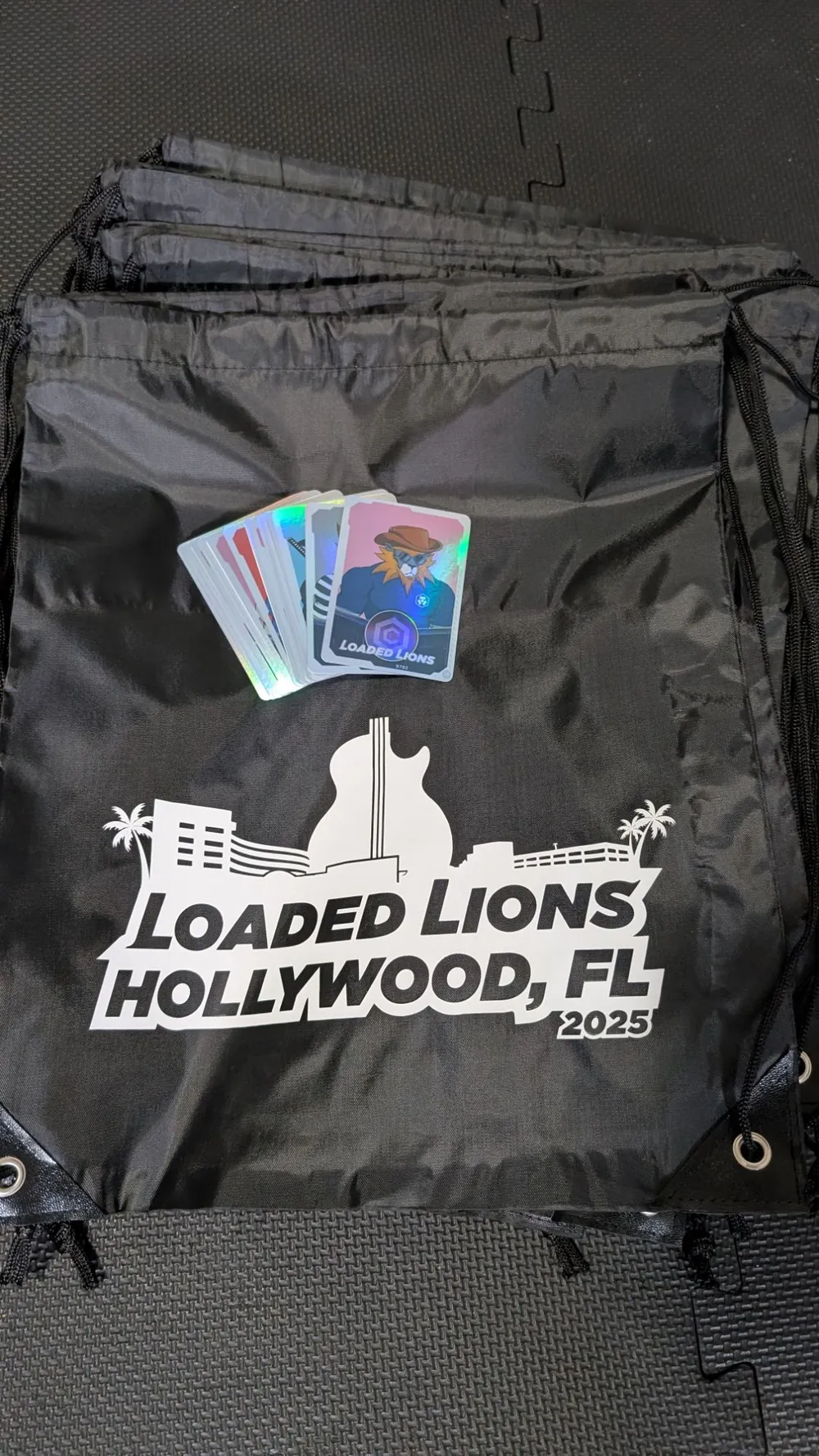 Black drawstring bags printed with 'Loaded Lions Hollywood, FL 2025' with holographic cards on top